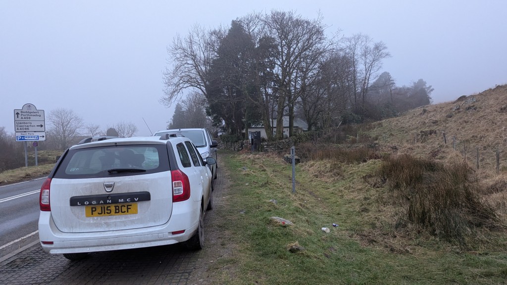 View of cars parked along the roadside near Pen y Gwryd, with a signpost in the background and fog obscuring the landscape.