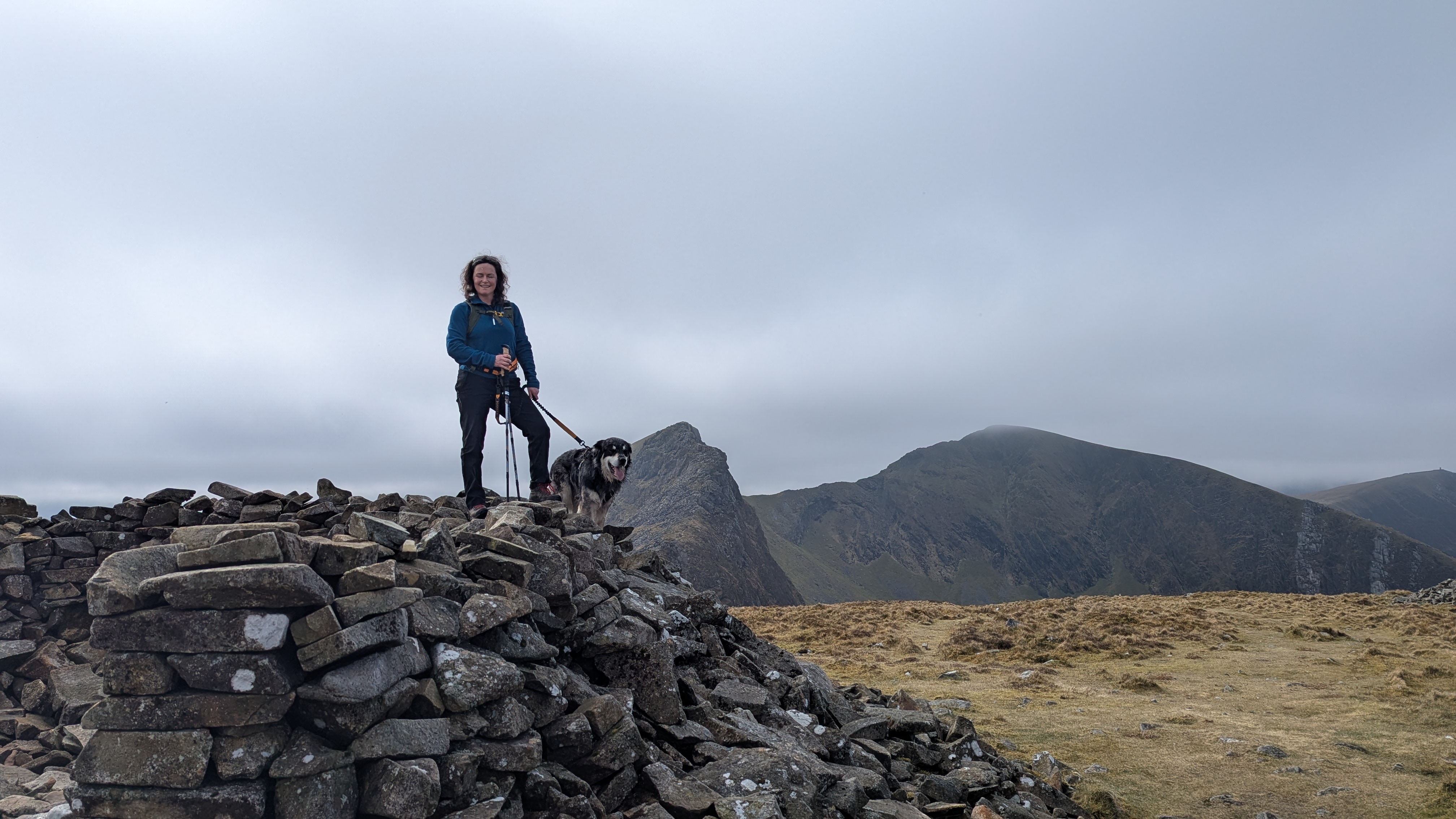 A person stands on a stone cairn with a dog, overlooking a mountainous landscape with overcast skies.