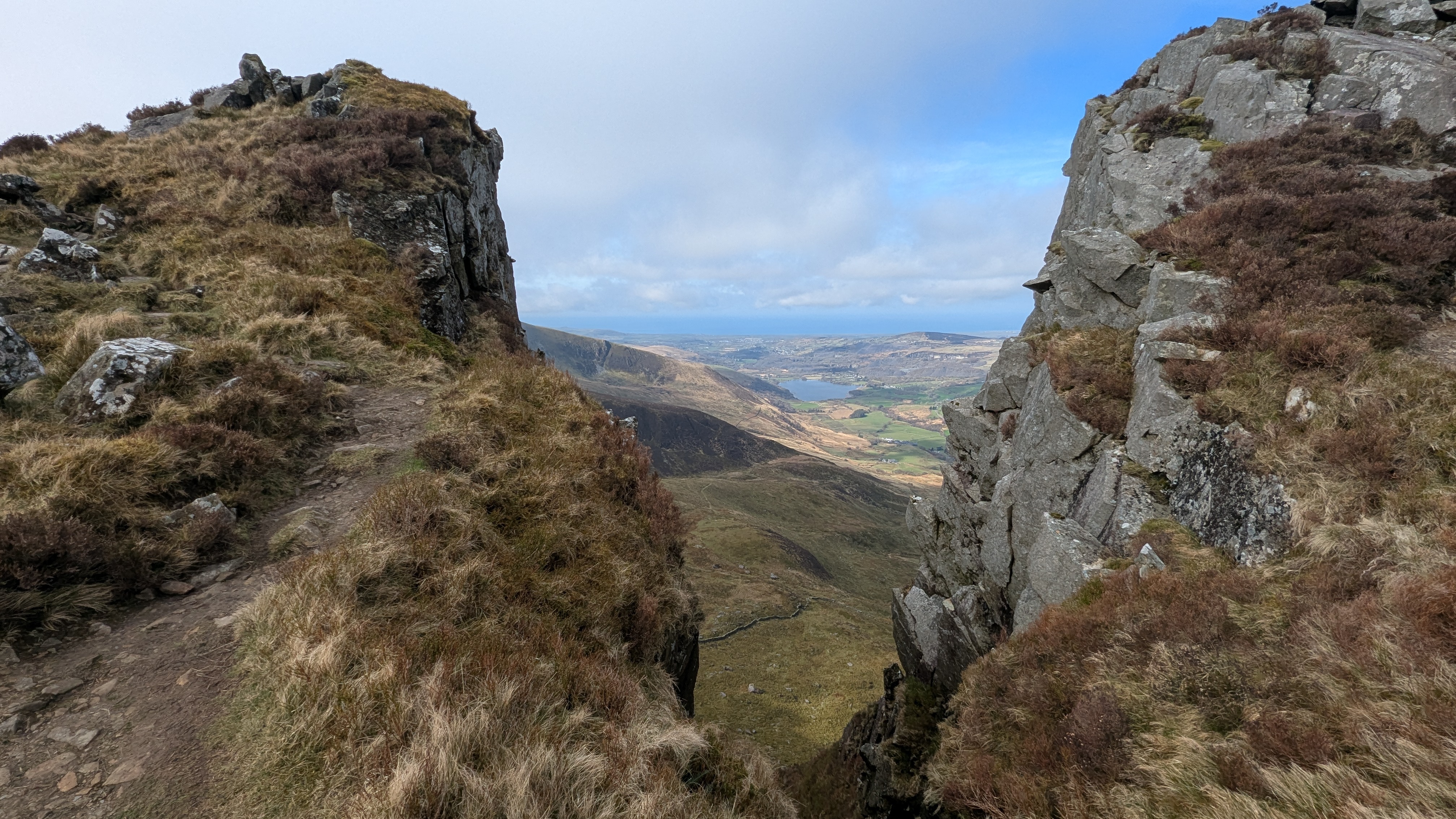 A narrow path between rocky outcrops on Nantlle Ridge, with views of valleys and hills in the background.