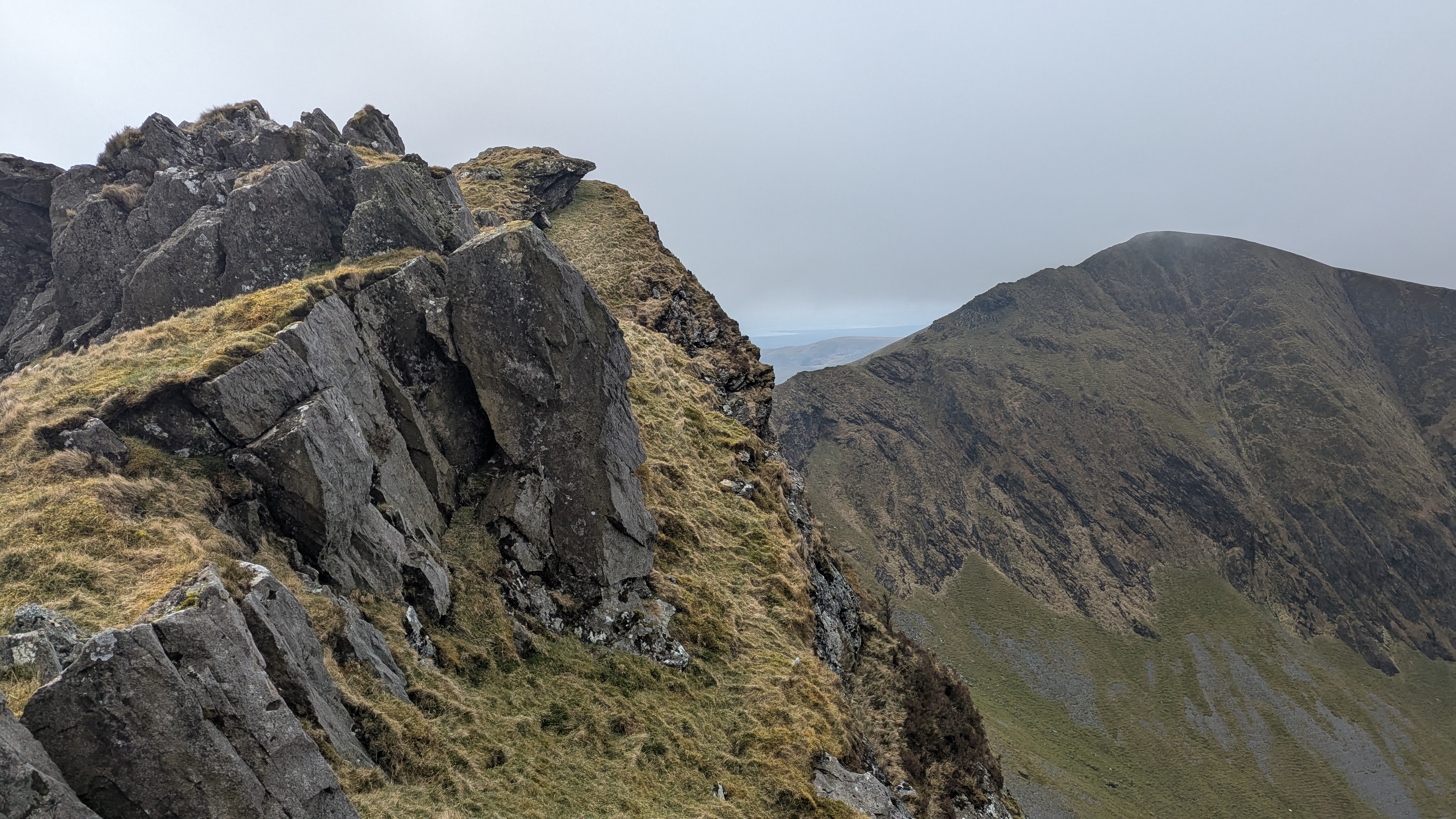 Rocky ridge with grassy edges and a panoramic mountain view in Eryri (Snowdonia), under overcast skies.