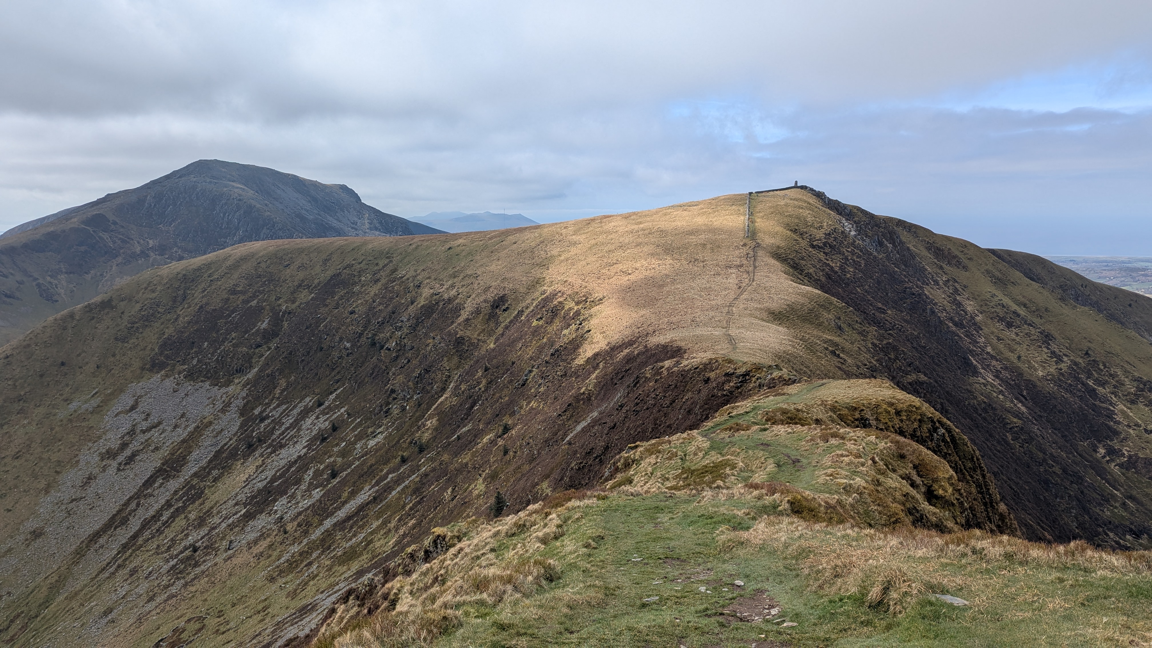 A panoramic view of Nantlle Ridge in Eryri (Snowdonia), showcasing grassy slopes and rugged terrain under a partly cloudy sky.