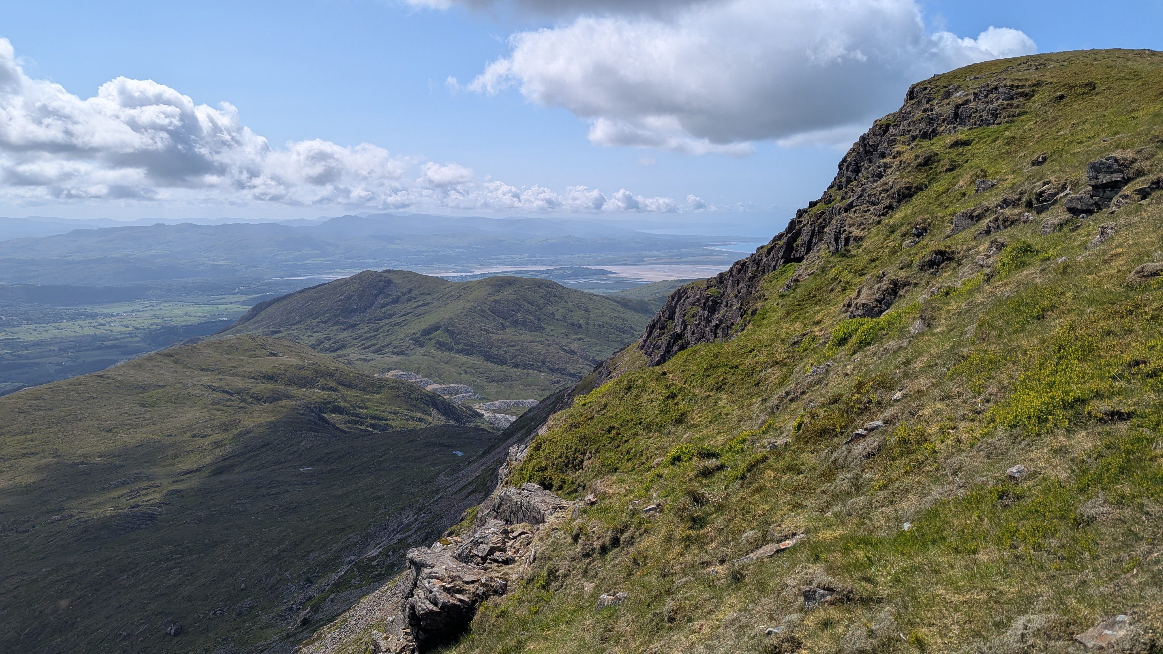 A panoramic view from the slopes of Moel Hebog, featuring green hills, rocky outcrops, and distant mountains under a partly cloudy sky.