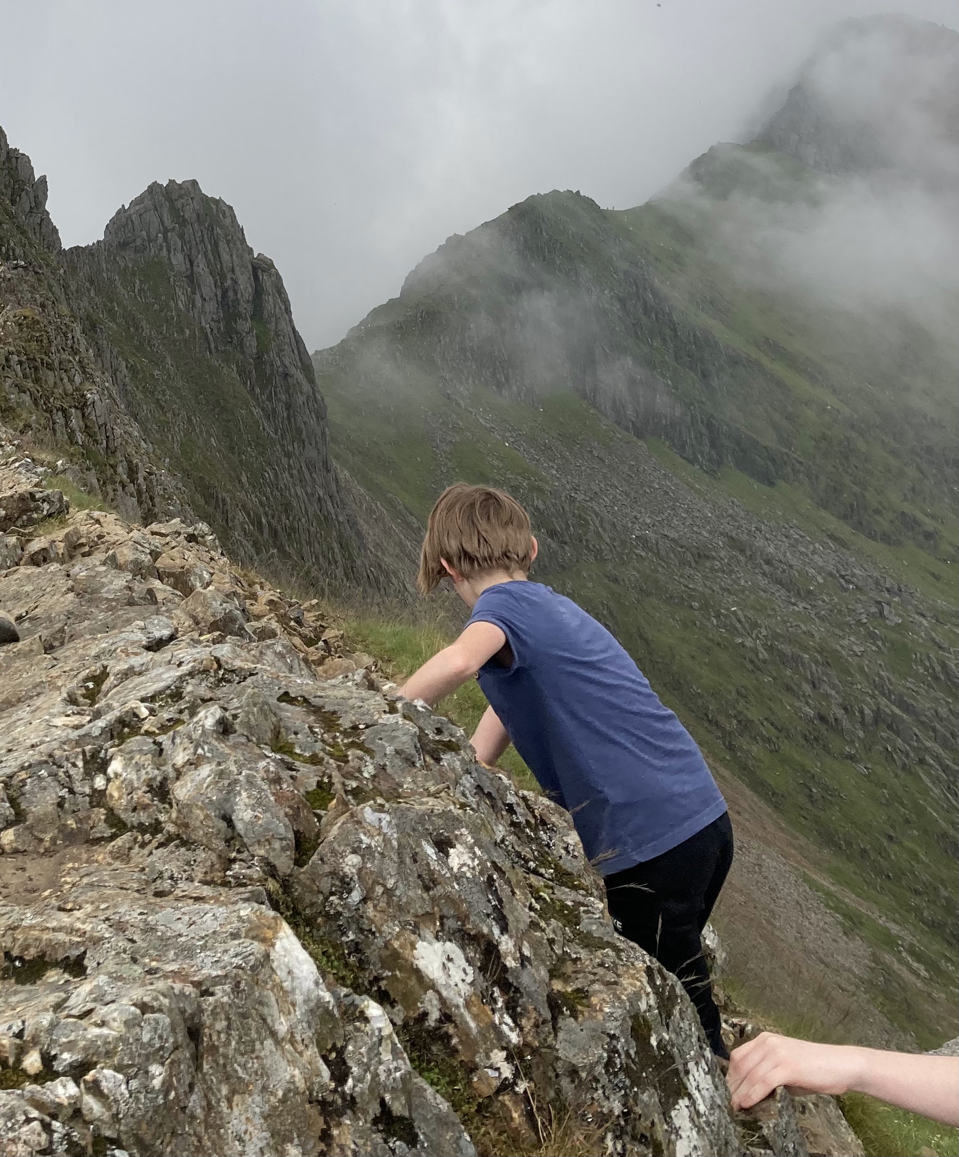 A child climbing a rocky hillside in a mountainous landscape, with fog and greenery visible in the background.