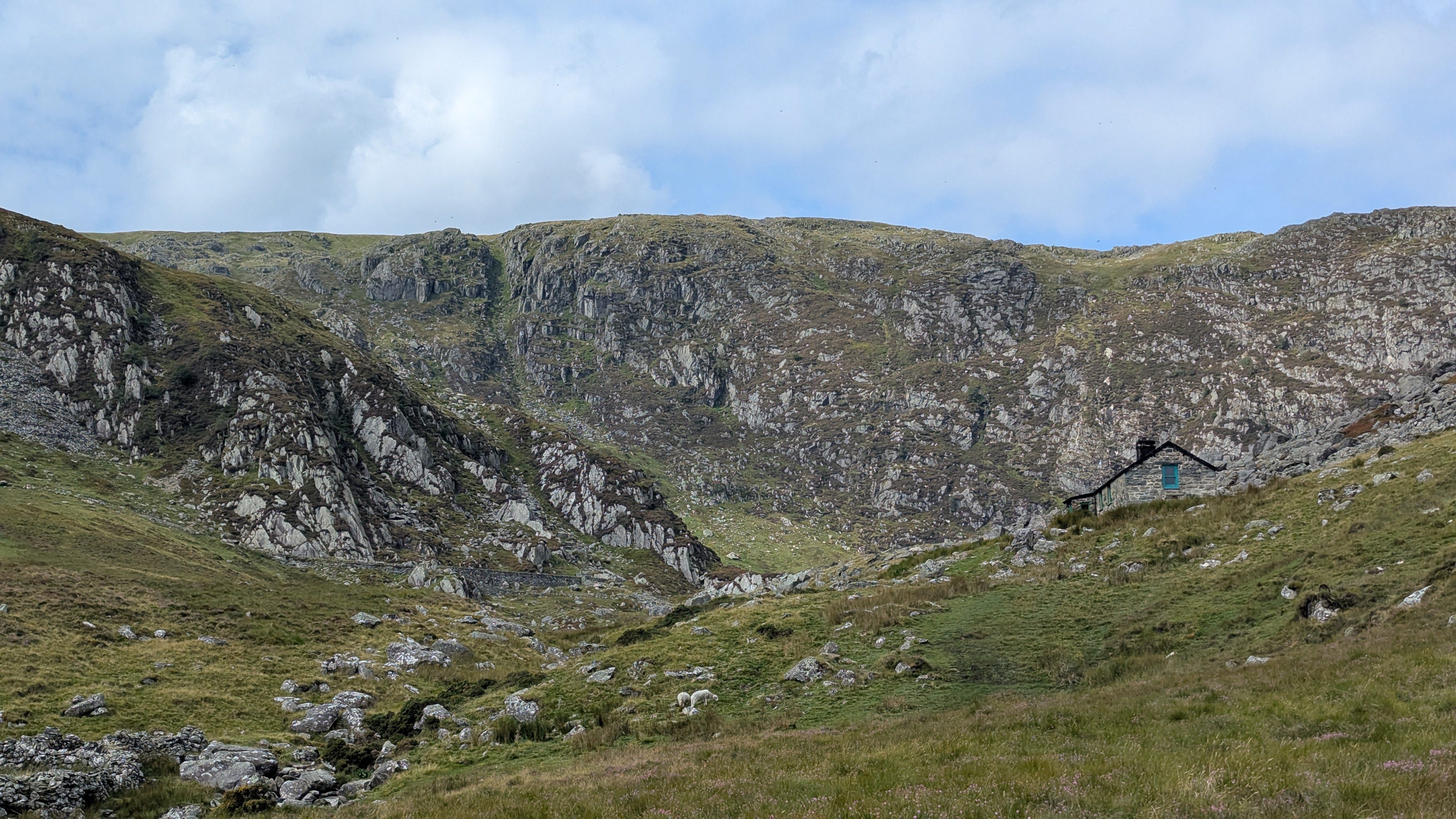 A picturesque view of a rugged valley in the Carneddau Mountain Range, featuring a small stone cottage surrounded by rocky terrain and grassland under a partly cloudy sky.