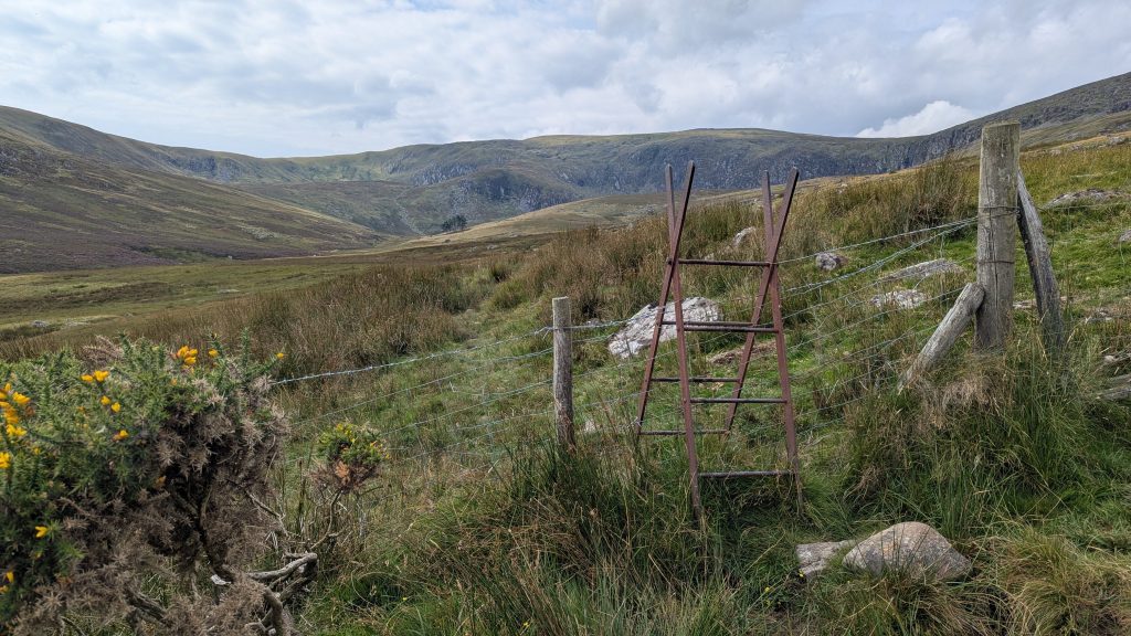 A scenic view of the Carneddau Mountain Range in Eryri, featuring a fence with a metal stile and a lush landscape with greenery and distant hills under a partly cloudy sky.
