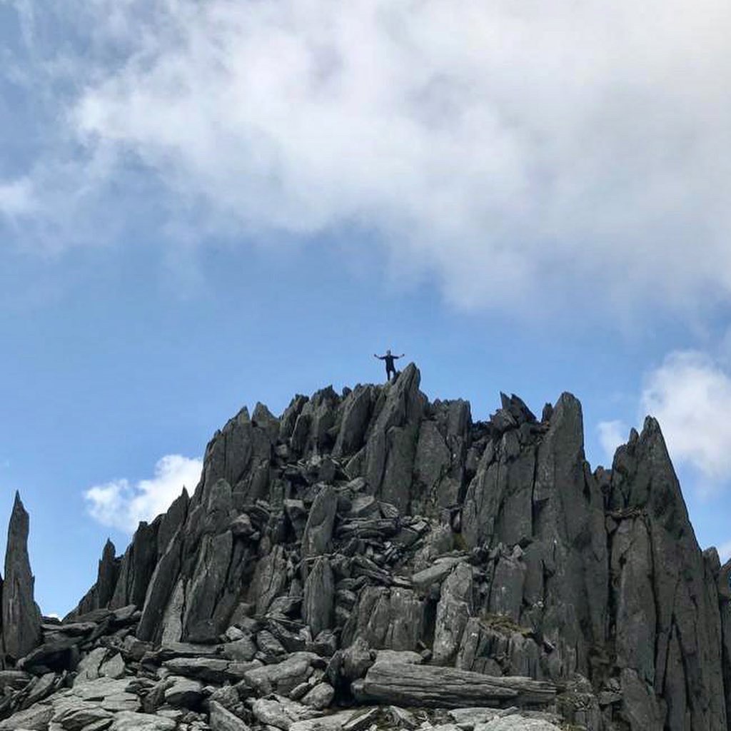 A climber stands triumphantly at the summit of a rocky outcrop, surrounded by jagged stone formations and a blue sky with scattered clouds.