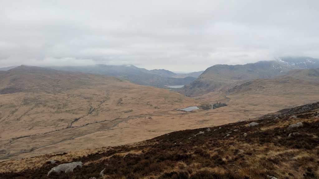 A panoramic view of the Glyderau mountain range, showcasing rolling hills and valleys under a cloudy sky, with a hint of snow on some peaks.