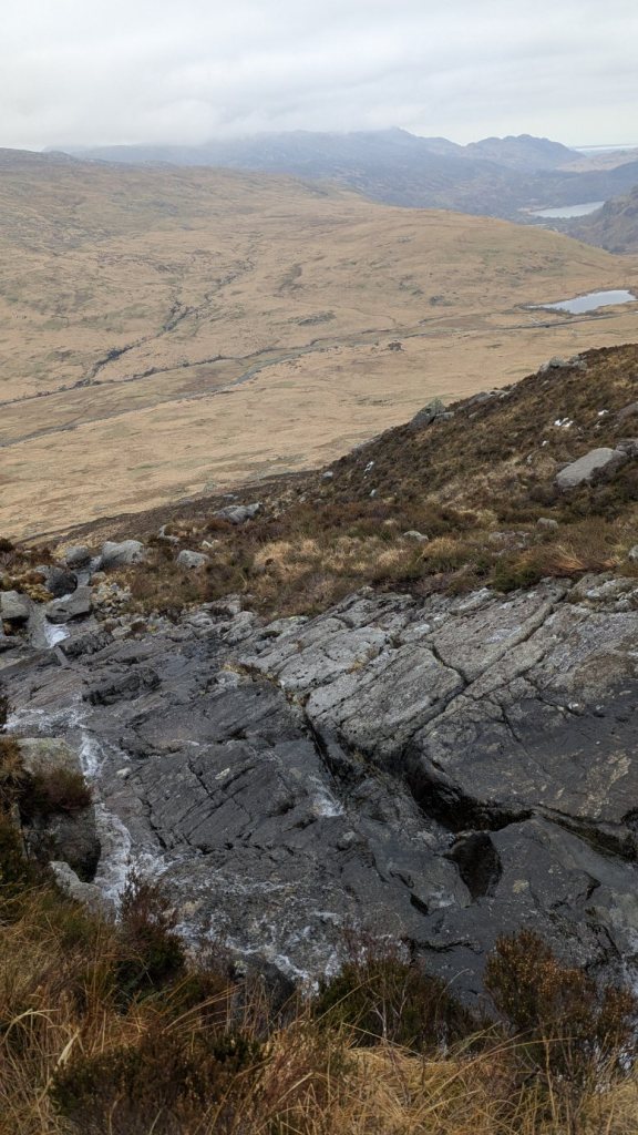 View of a rocky landscape with a stream flowing down a slope, surrounded by heather and grass, in the Glyderau mountains.