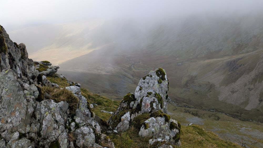 View from the rocky ridge overlooking a misty valley, with grassy slopes leading down to a river.