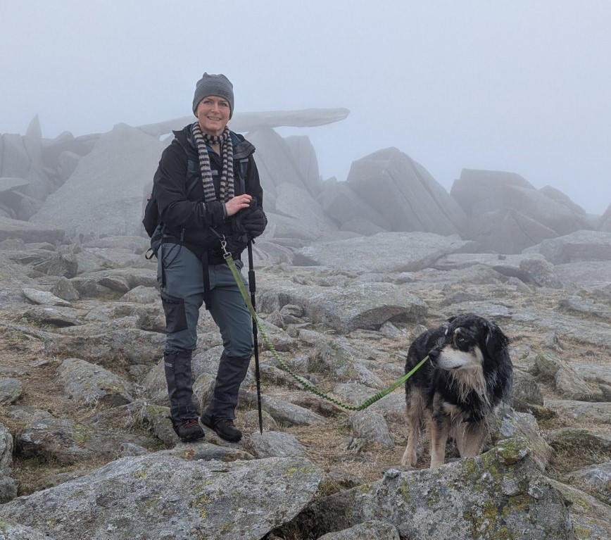 A person standing on rocky terrain in misty conditions, holding a walking stick with a dog beside them. The background features large rock formations partially obscured by fog.