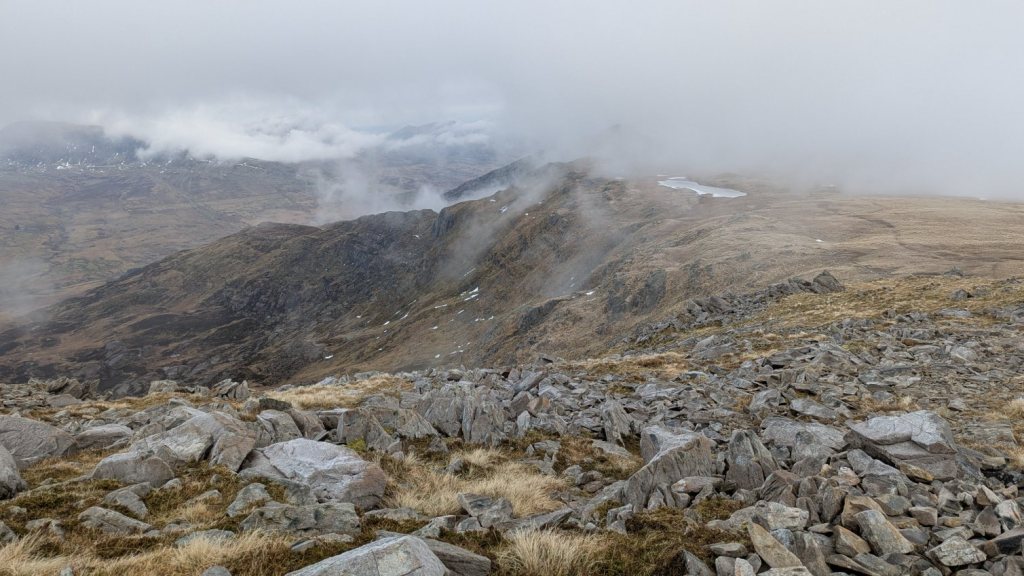 A rocky mountainous landscape shrouded in clouds, with patches of grass and snow visible on the slopes.
