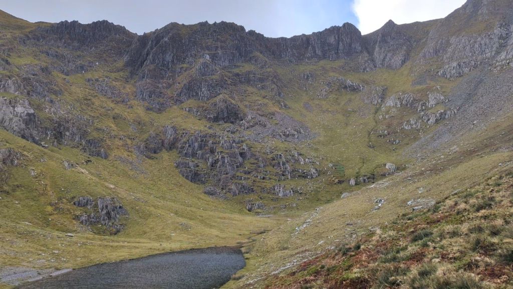 View of the rugged landscape leading to Yr Elen, showcasing steep rock formations and a small body of water in the foreground, surrounded by grassy slopes.