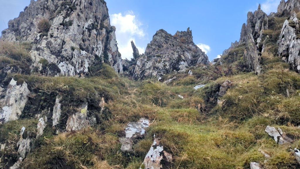 View of the Dragon's Teeth Ridge, a rocky and spiky terrain on the ascent to Yr Elen, with grass-covered slopes and a blue sky.