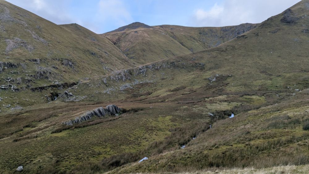 A wide valley view of the Carneddau mountain range, featuring grassy slopes and rocky outcrops under a cloudy sky.