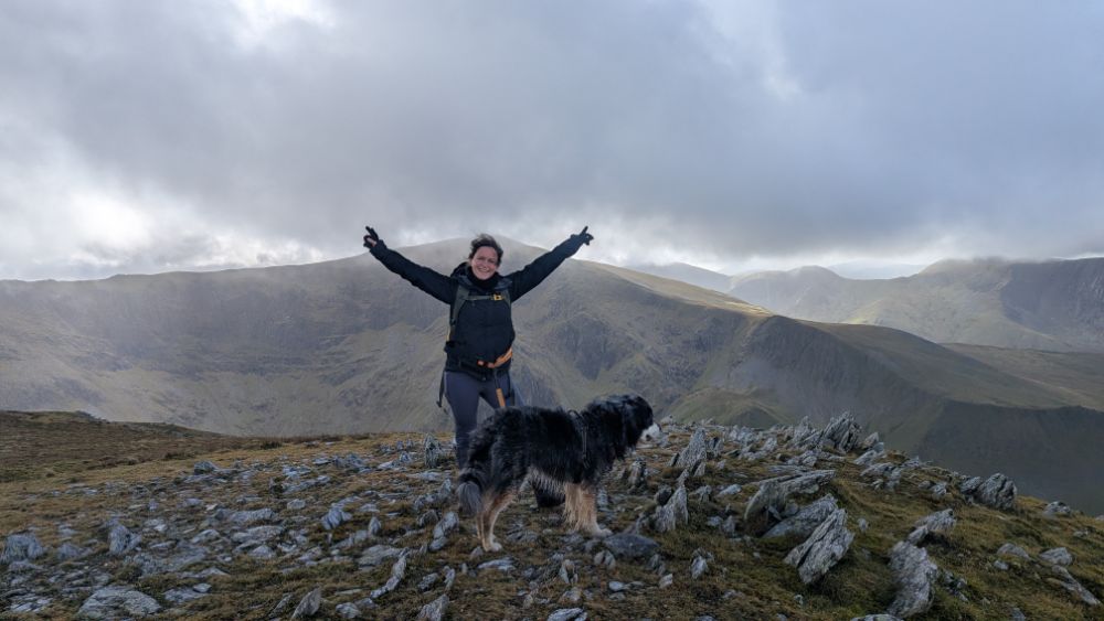 Hilary Pullen with outstretched arms stands on a rocky mountain ridge alongside her dog Ziggy, with a cloudy mountainous landscape in the background.