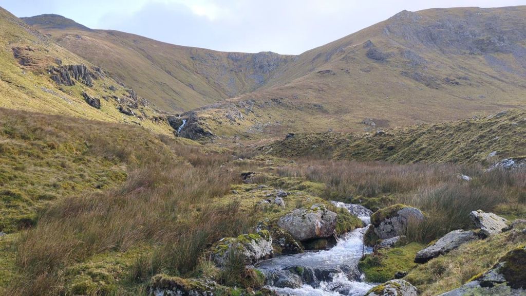 A scenic view of a valley with rocky terrain, grass, and a small stream flowing through it, framed by mountainous slopes in the background.