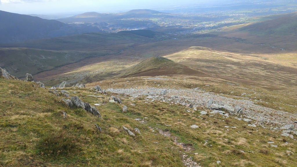 A panoramic view from Yr Elen's summit, showcasing rolling hills and valleys of the Carneddau mountain range with patches of grass and rocky terrain.