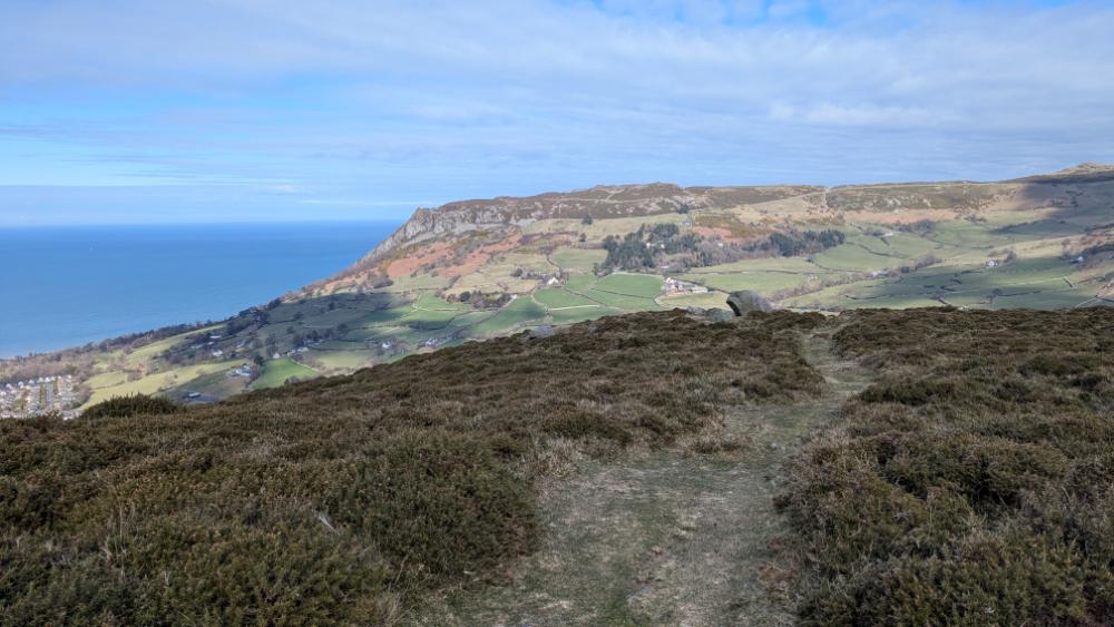A panoramic view of a hilly landscape with lush green fields and a coastline by the sea under a partly cloudy sky.