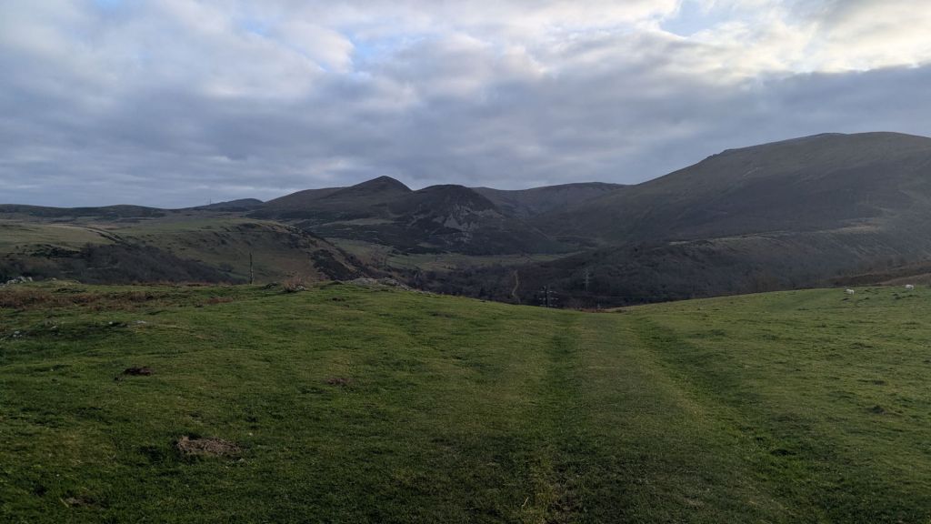 View of the rolling hills and mountains surrounding Bera Bach in the Carneddau, with a grassy path leading into the distance under a cloudy sky.