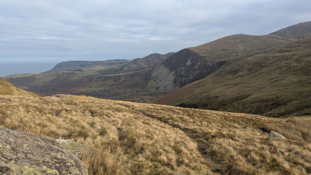 A panoramic view of the Carneddau mountains and the North Wales coastline, showcasing grassy slopes and rolling hills under a cloudy sky.