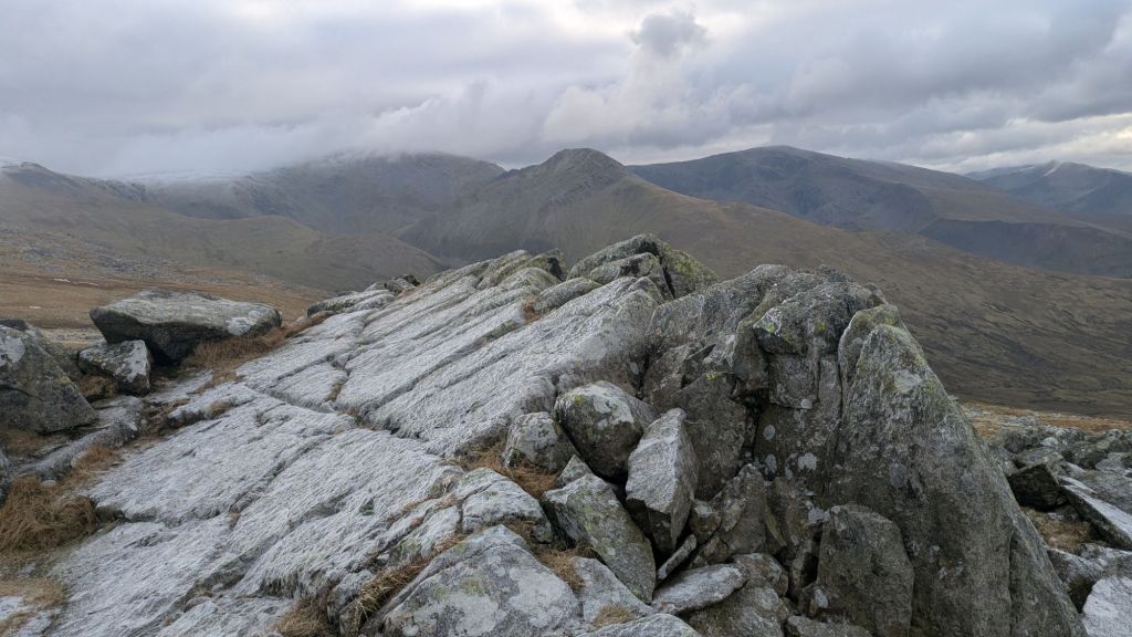 A rocky summit of Bera Bach, with large stone formations in the foreground and distant mountain ranges in the background, under a partly cloudy sky.
