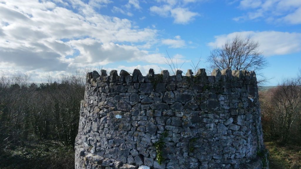 Aerial view of an ancient stone tower with a crenellated top, surrounded by trees and a blue sky with clouds.