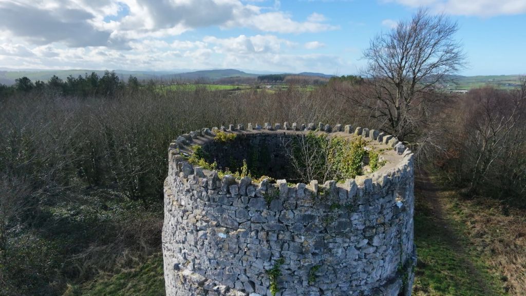 An aerial view of a stone tower ruin surrounded by trees, with blue skies and rolling hills in the background.
