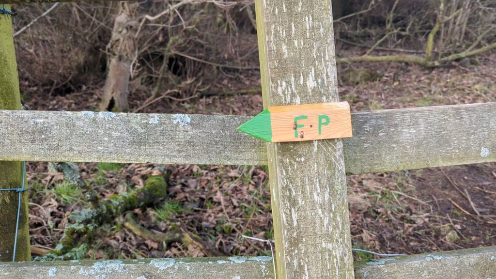 A wooden fence post with a directional sign painted in orange and green, indicating 'F.P.' with an arrow pointing to the right, set in a natural outdoor environment.