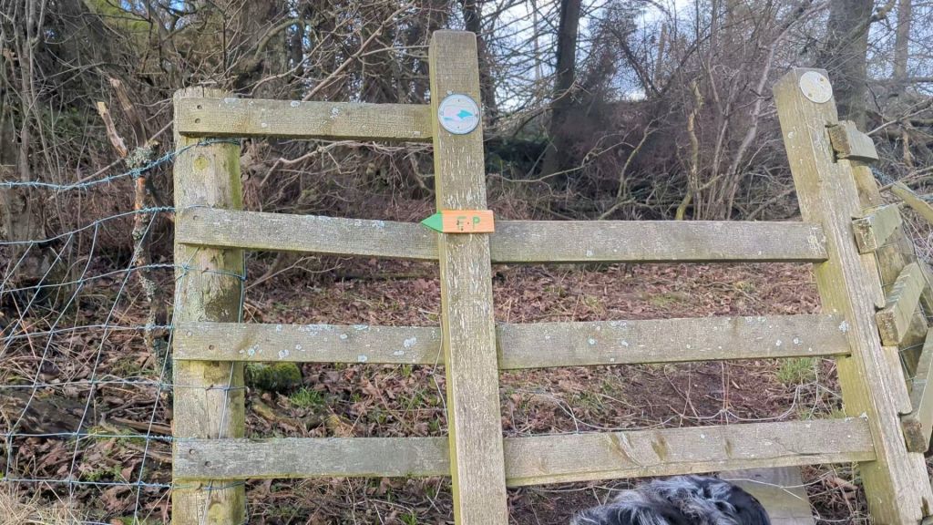 A wooden gate in a rural area with a 'Footpath' sign and two circular markers attached, surrounded by trees and underbrush.