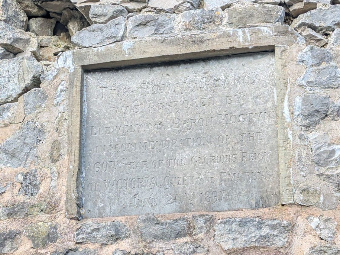 A weathered stone wall featuring an engraved plaque commemorating the 50th year of Queen Victoria's reign, surrounded by natural elements and a metal grate.