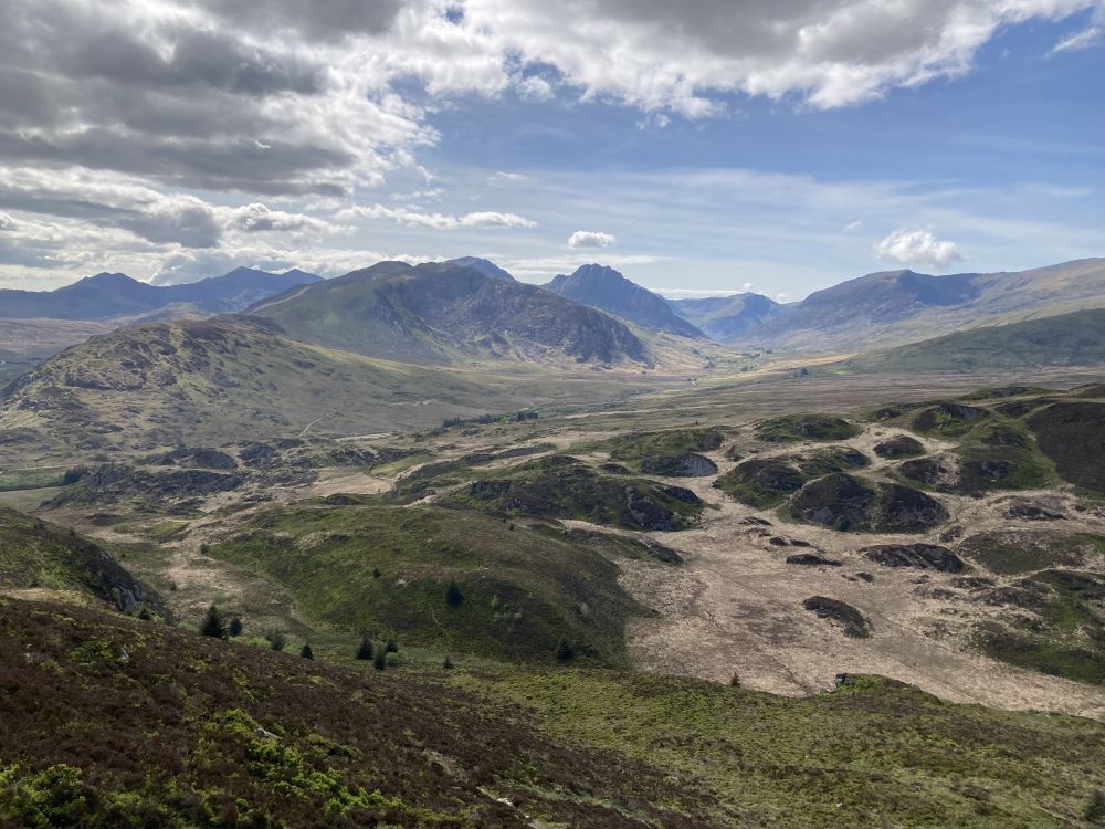 A panoramic view of a rugged landscape featuring rolling hills and mountains under a blue sky with scattered clouds.