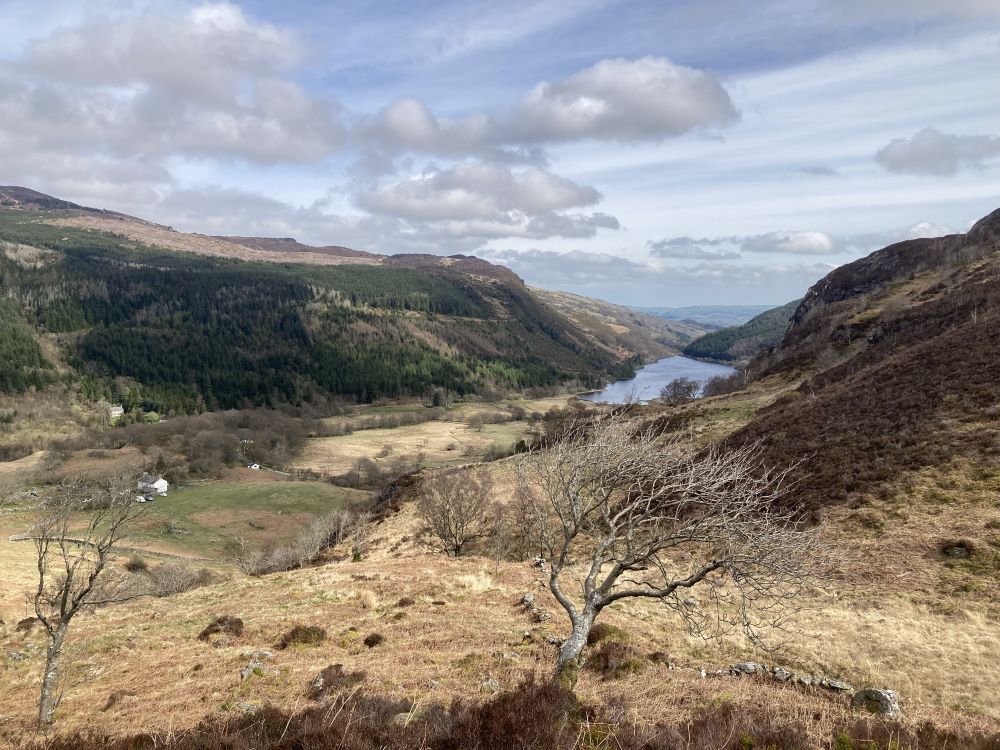 A scenic view of a valley with a river running through it, surrounded by rolling hills and forests under a partly cloudy sky.