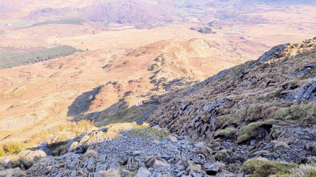 A scenic view from a high vantage point overlooking rugged terrain and rolling hills, with a mix of grass and rocky surfaces in the foreground and valleys stretching into the distance.