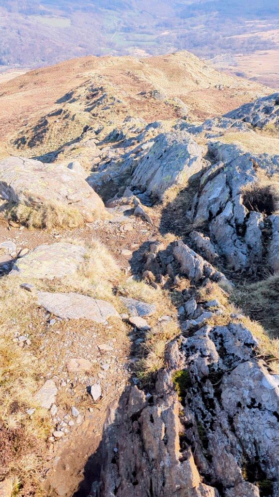 A rocky trail winding through a mountainous landscape, with grass and boulders on either side and a view of distant hills in the background.