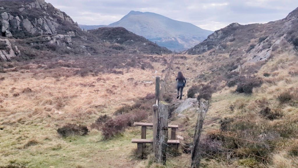 A hiker walking along a narrow path through a grassy valley surrounded by rocky hills and mountains under a cloudy sky.