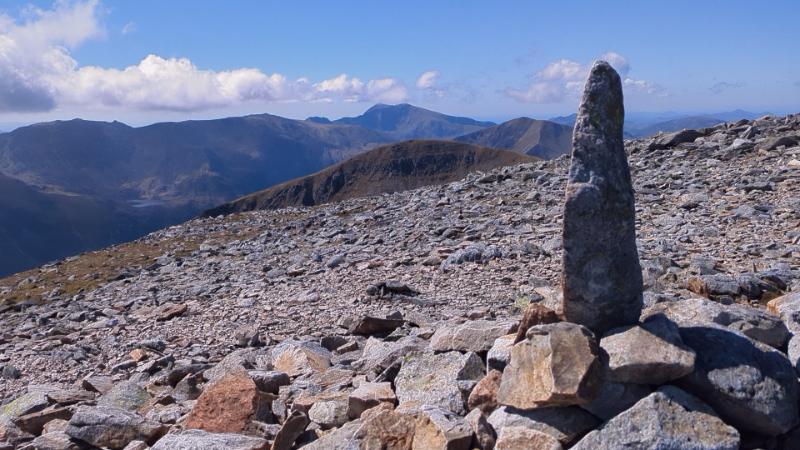 A tall stone monolith standing amidst a rocky landscape with distant mountains under a clear blue sky.