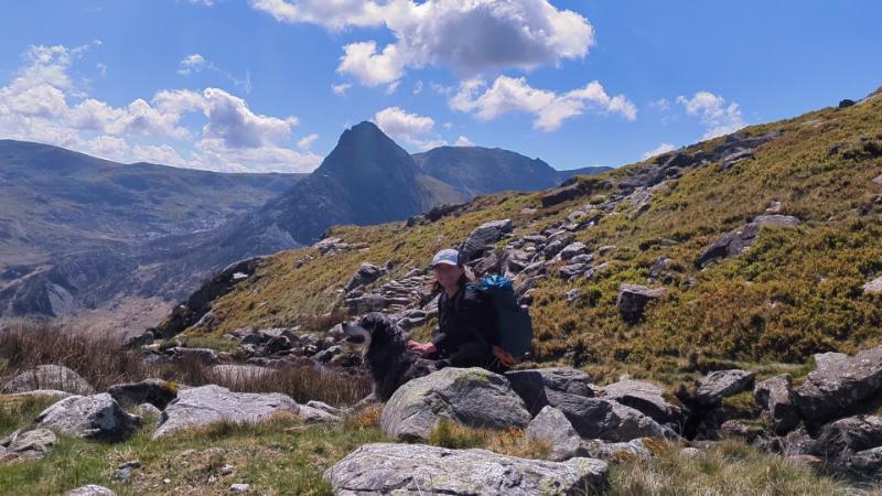 A person sitting on rocks with a black dog in a mountainous landscape under a blue sky with clouds.
