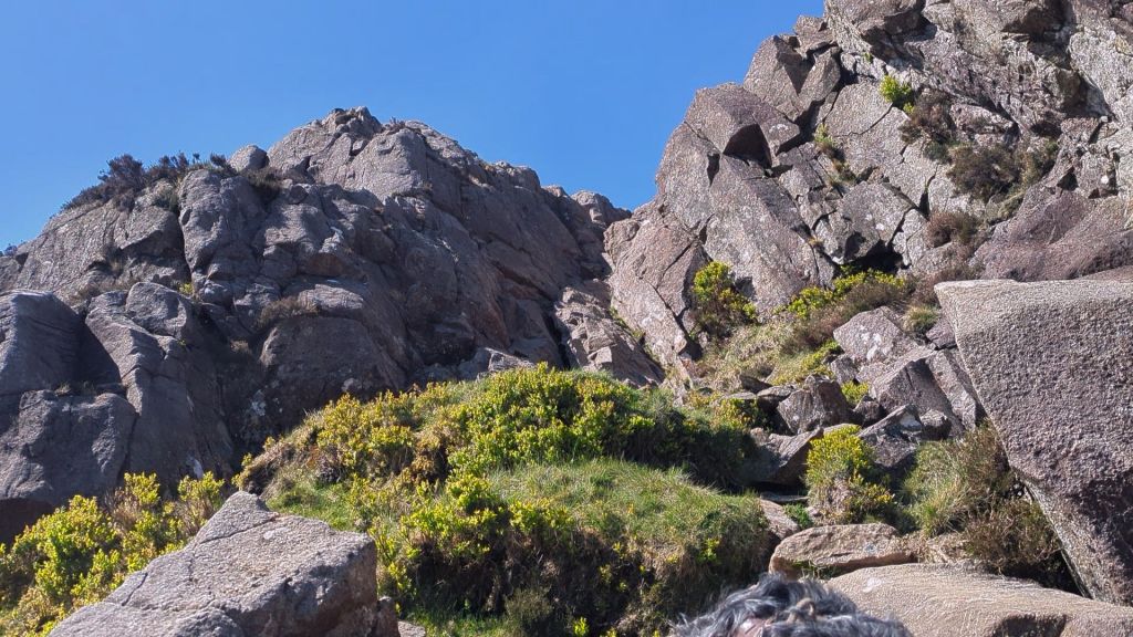 A view looking up at rocky terrain and cliffs under a bright blue sky, with patches of green grass and shrubs in the foreground.