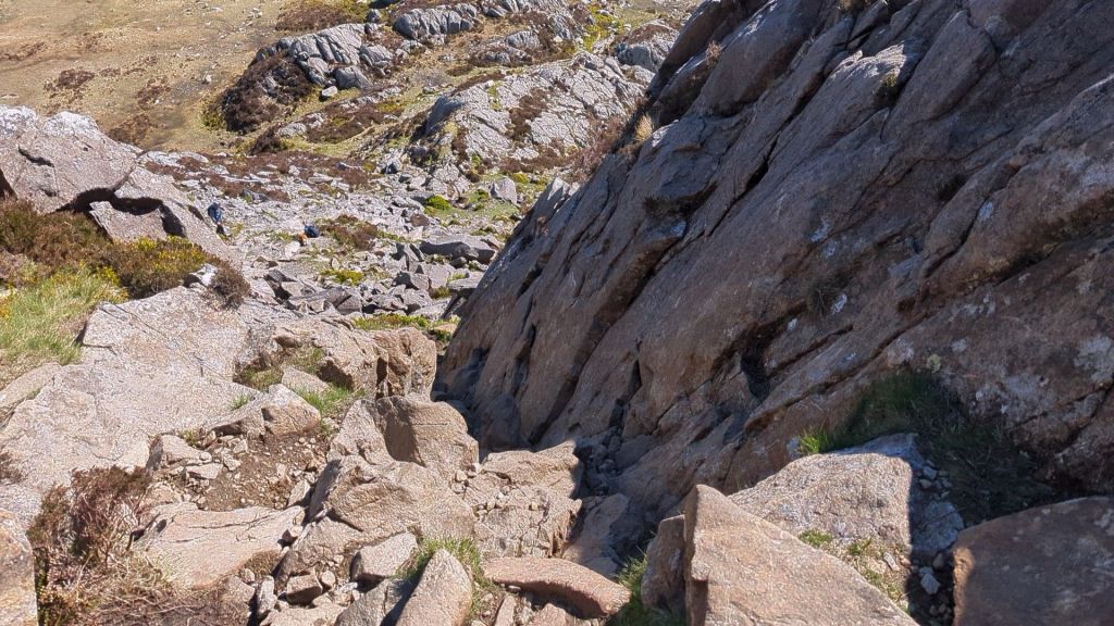 A steep rocky hillside with loose stones and sparse vegetation, showing a rugged landscape in natural light.