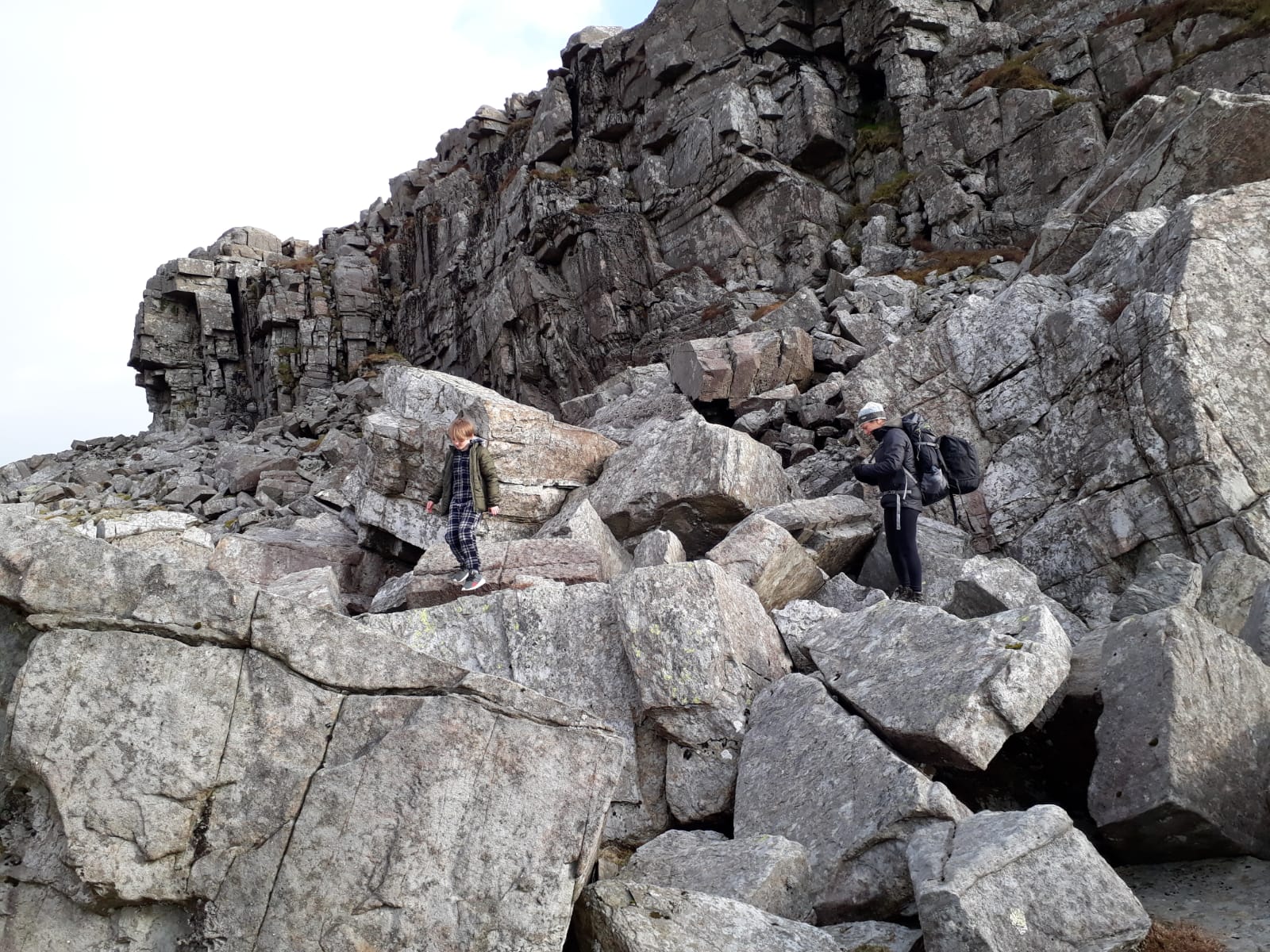 A child carefully navigates large rocky terrain while an adult with a backpack stands nearby, surrounded by steep rock formations.