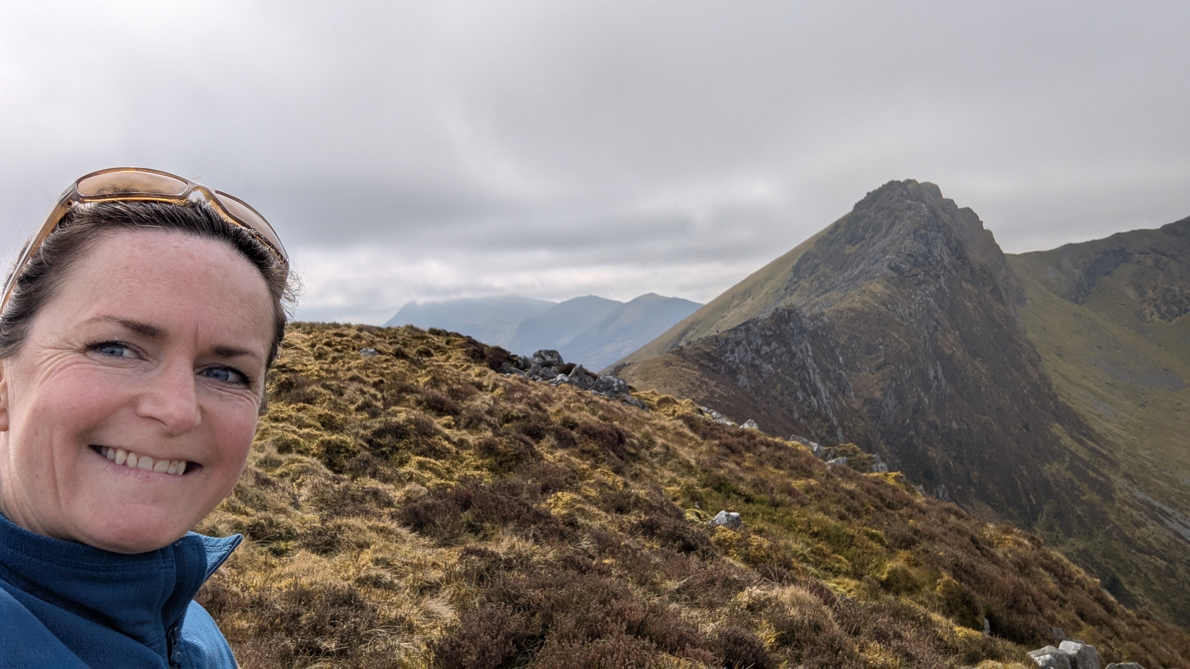 A smiling woman taking a selfie on a mountainous landscape, with rolling hills and rocky peaks in the background under a cloudy sky.