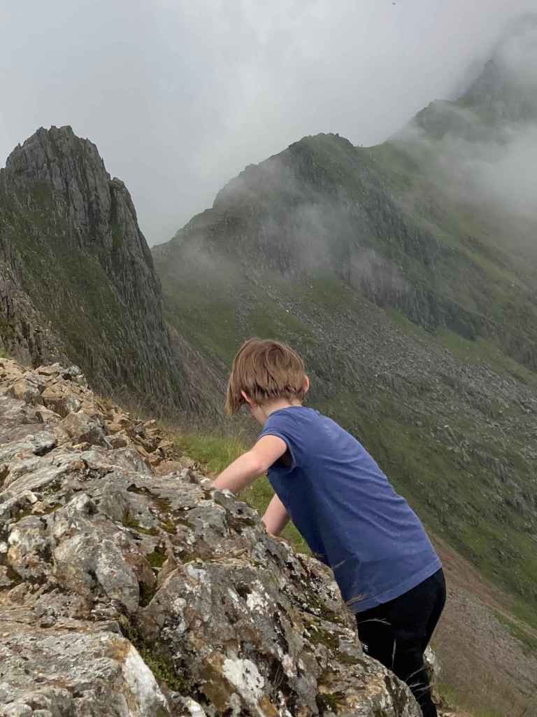 A child climbing a rocky mountain trail surrounded by misty peaks and clouds.